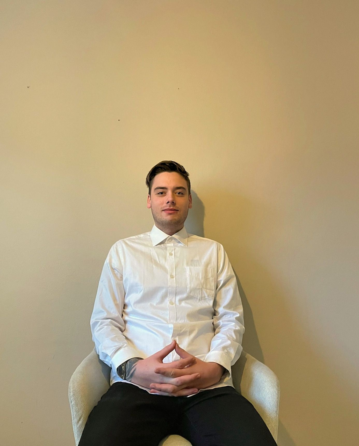 Person sitting in a chair against a plain beige wall, wearing a white shirt and black pants.